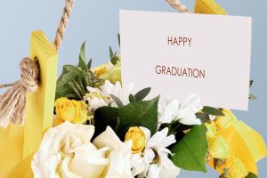 A beautiful bouquet filled with white and yellow flowers hangs against a soft blue background. a card proudly displays the words happy graduation, perfect for celebrating this special day.